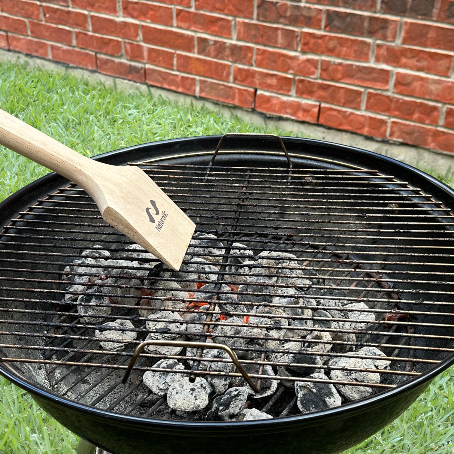 Wooden Grill Scraper 14” resting on a barbecue grill with charcoal, showcasing its bristle-free cleaning design.